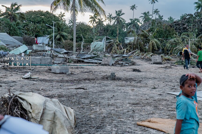 Young boy in blue tee looks to camera as men work behind him on land devasted by a tsunami, Tonga. 
