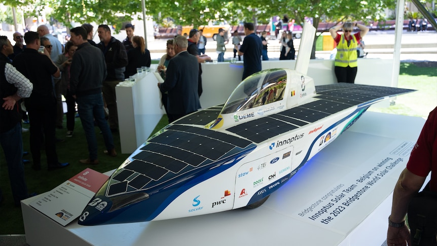 A sleek racing car covered in solar panels sits on a raised podium.