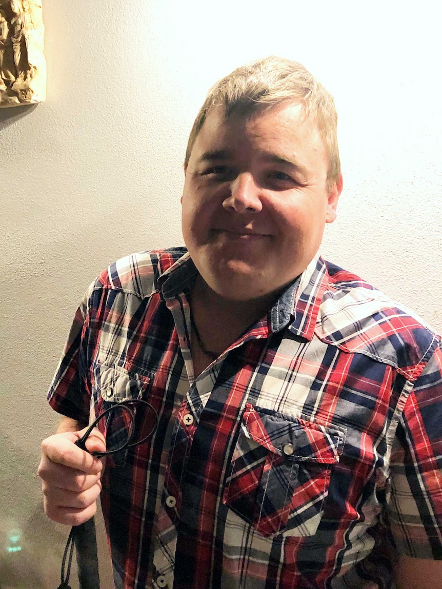 Conor Smith, dressed in a plaid shirt, stands in front of a white wall and holds a dog leash.