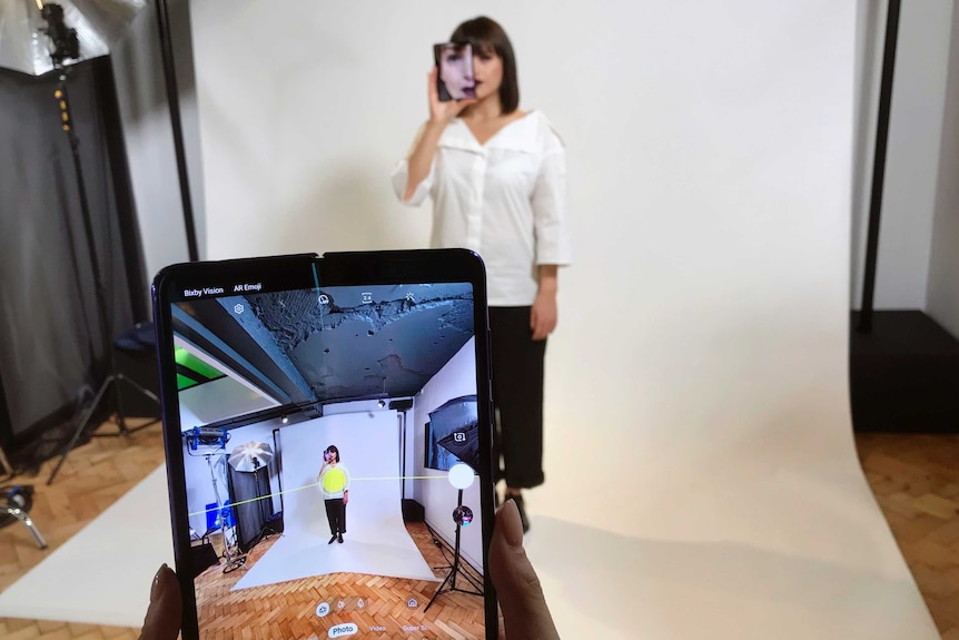 A foldable phone is held up to film a model holding up a mobile device to her face in front of a white backdrop