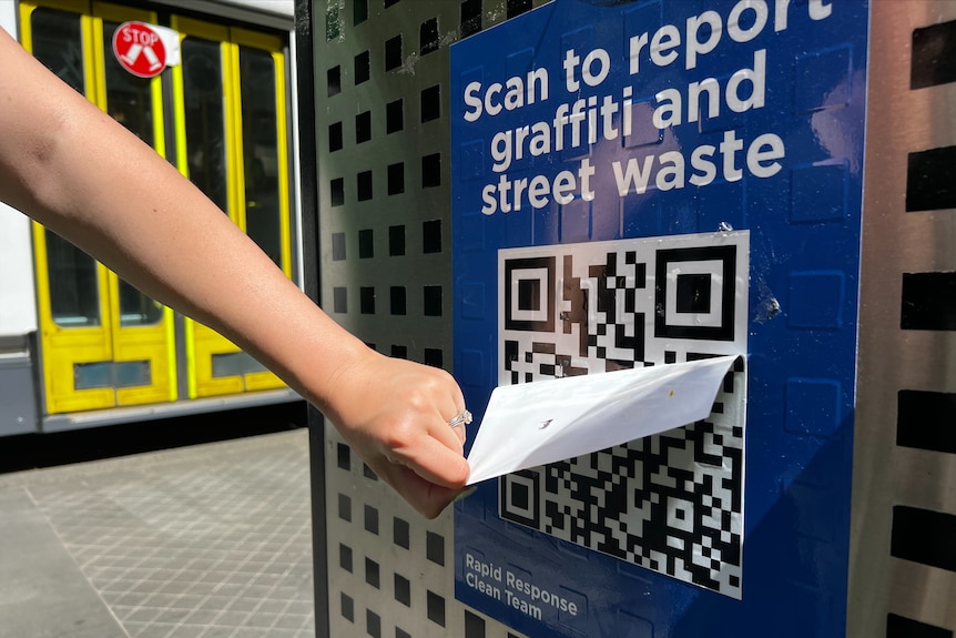 A hand tearing a fake QR code off a poster bearing a genuine QR code to a graffiti reporting system.