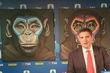 A man in a suit speaks in front of three paintings of apes.