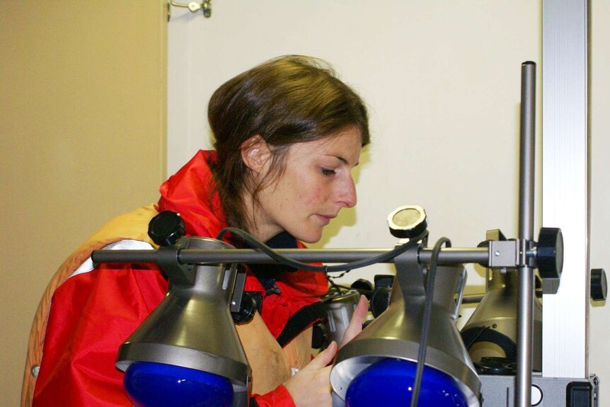 Profile of woman in a large red jacket, long brown hair surrounded by scientific equipment. 2 lights with blue bulbs in the fore