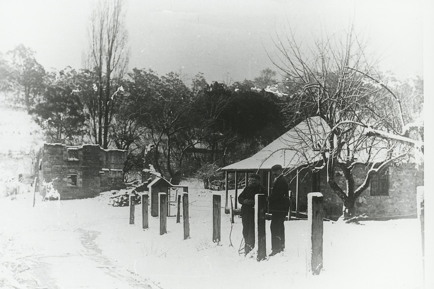 Black and white photograph, two men stand in a snow filled scene complete with cute snow covered cottage