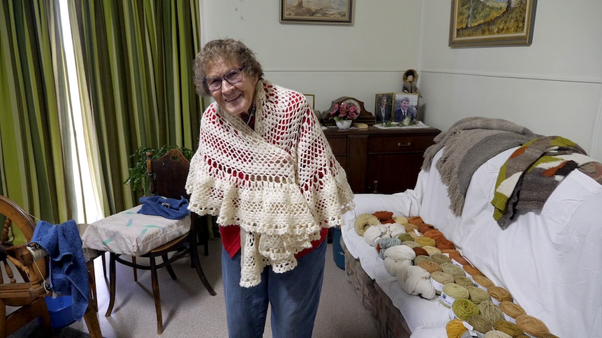 An elderly woman wearing a white knitted shawl. She stands next to a couch laden with wool.