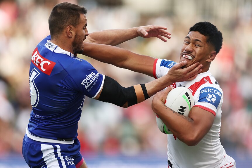 A St George Illawarra NRL player fends off a Canterbury opponent with his right hand while holding the ball with his left.