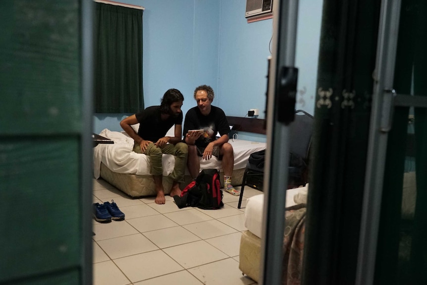 Two men sit on a bed, looking down at a device.