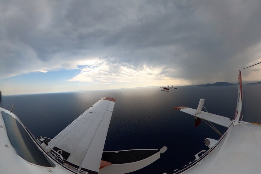 Planes fly over water with a large plume of smoke in the background.