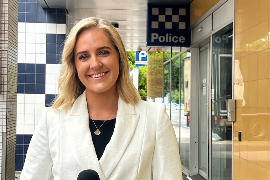 A blond woman wearing a blazer stands in front of a police station 