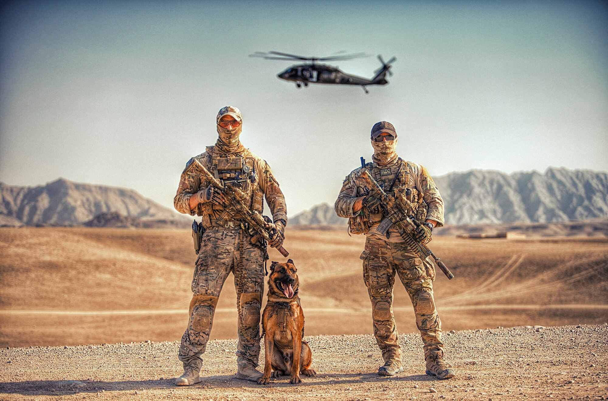 Two soldiers holding weapons pose in the desert with a dog and a helicopter hovering behind them