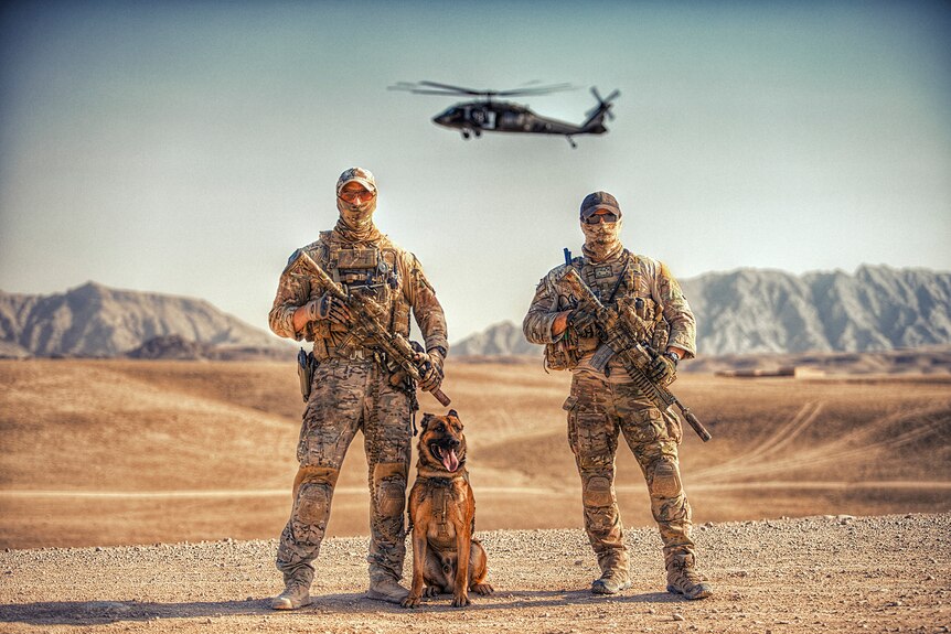 Two soldiers holding weapons pose in the desert with a dog and a helicopter hovering behind them