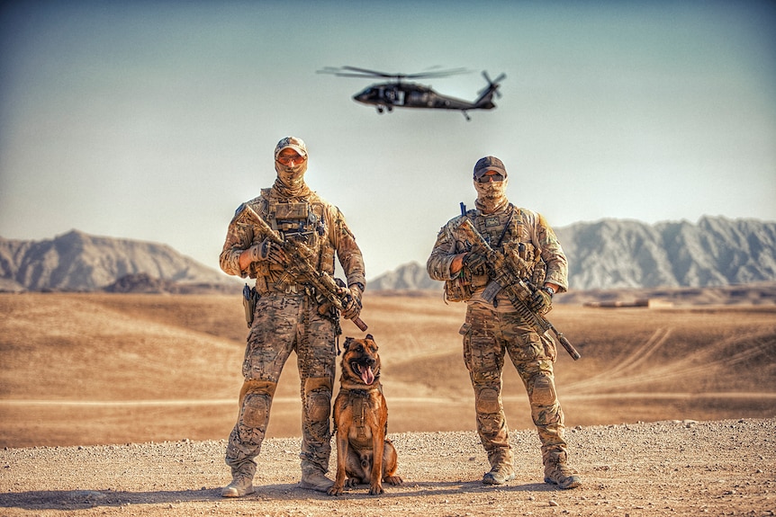 Two soldiers holding weapons pose in the desert with a dog and a helicopter hovering behind them
