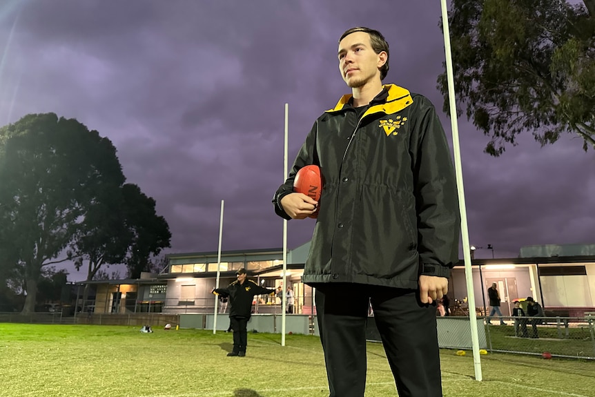 A stoic football club secretary stands on a sprawling AFL field during twilight