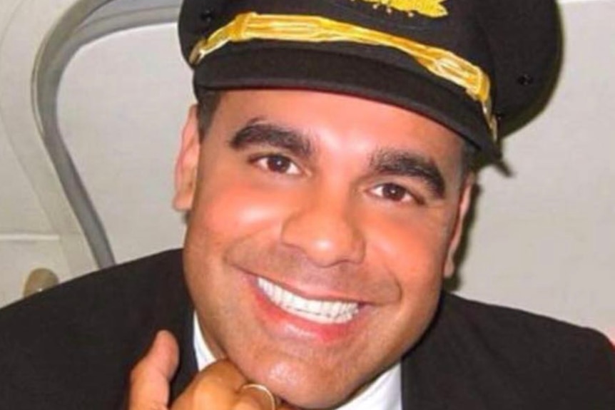 A man in a flight attendant's suit and cap smiles at the camera from inside an airplane cabin, he deals with stressed passengers