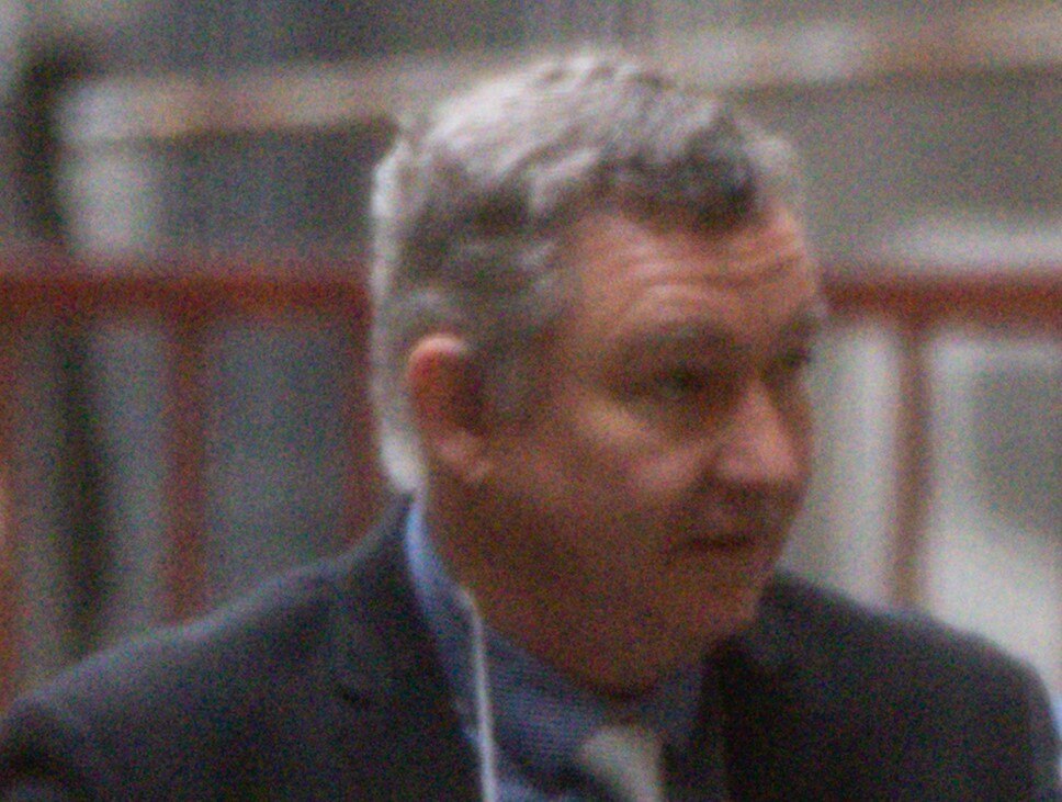 Greg Lynn wearing a suit and glasses around his neck being guided into court by a police officer.