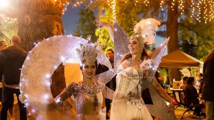 Two female dancers in elaborate silver costumes to represent a crescent moon and a star pose in a park at night.