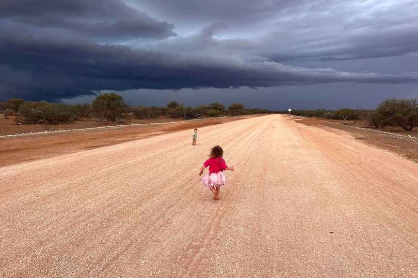 A young child wearing pink walks on a road barefoot before a dark storm