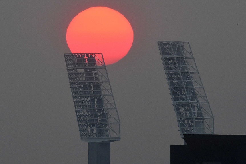 A sun rises into a grey sky over two light towers at the WACA Ground in Perth.