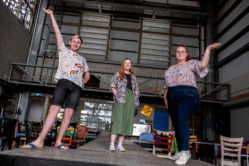 Three young people stand on a stage in a warehouse space.
