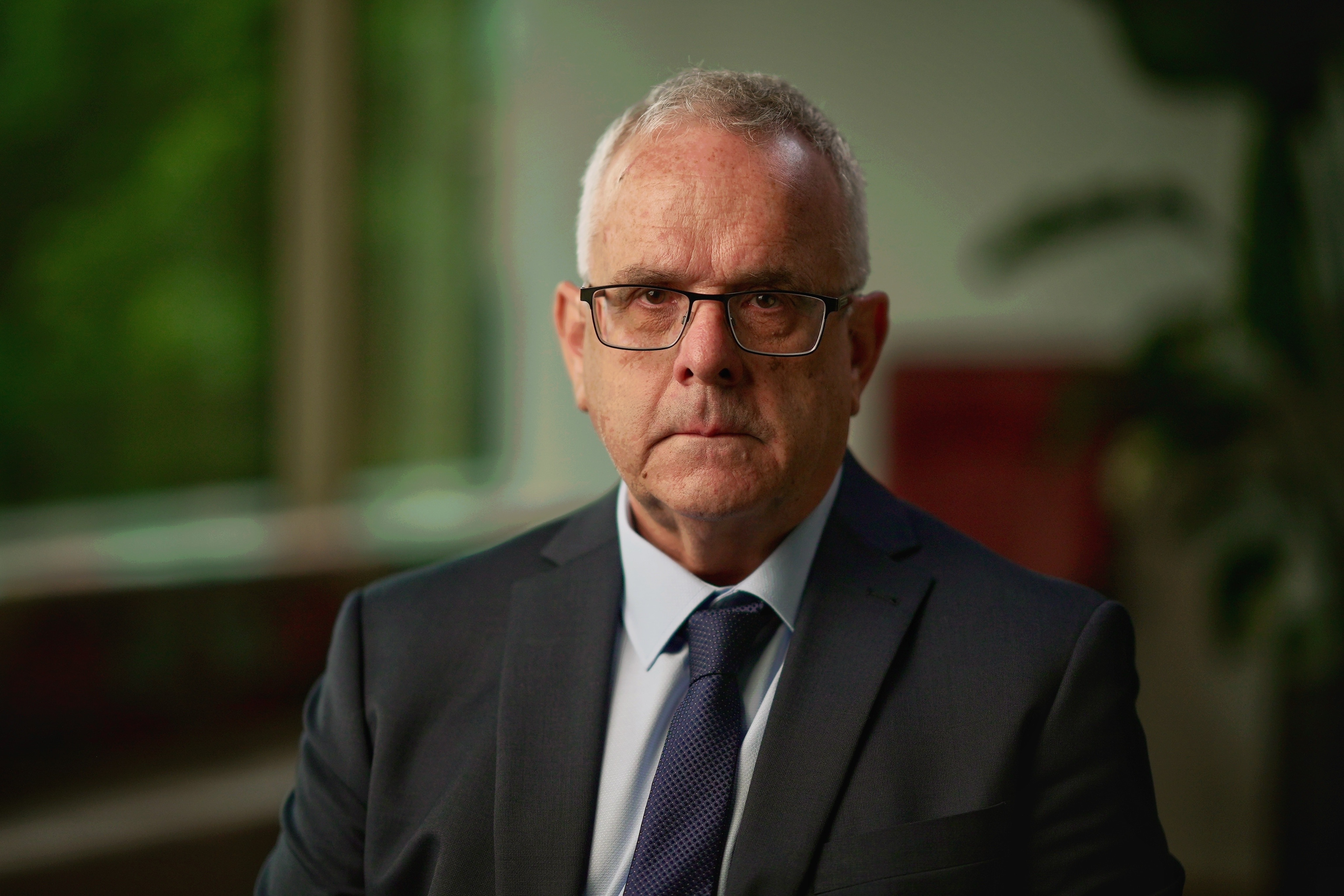 A man with grey hair and glasses, dressed in grey suit and tie, looks sternly at camera.