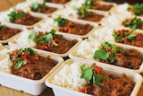 Containers with goat curry and rice topped with fresh coriander and chilli lined up on a benchtop