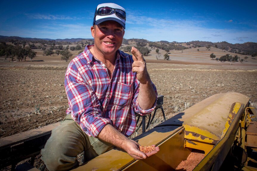 A farmer sitting on a plant seeder in a dry paddock smiling at the camera with his fingers crossed
