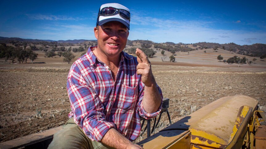 A farmer sitting on a plant seeder in a dry paddock smiling at the camera with his fingers crossed