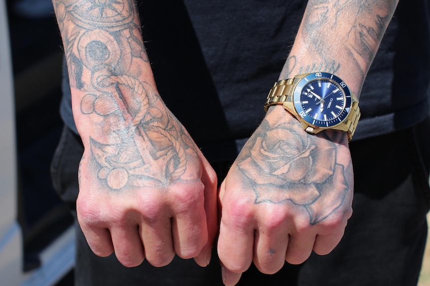Two tattooed hands with fists closed held out to camera, gold wrist watch, anchor tattoo on one had and and a rose on the other 