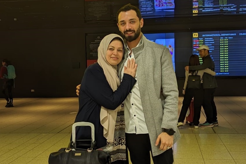 A mother and son stand with a suitcase under a "Departures" sign in an airport.