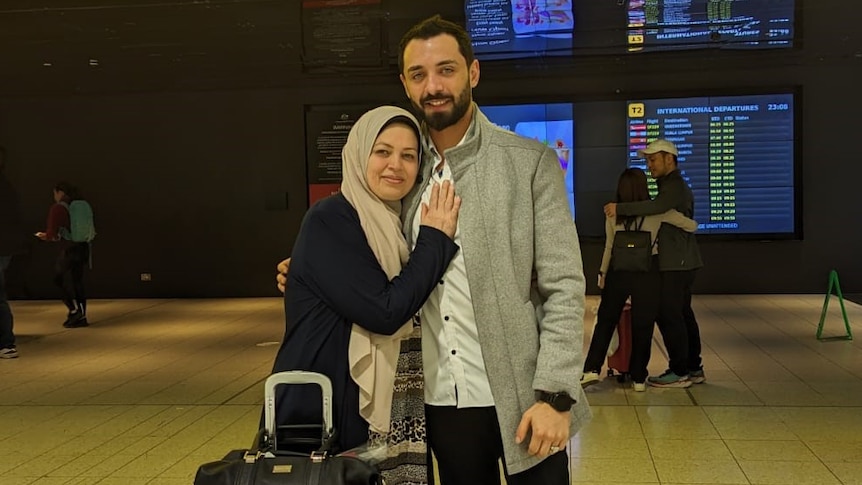 A mother and son stand with a suitcase under a "Departures" sign in an airport.