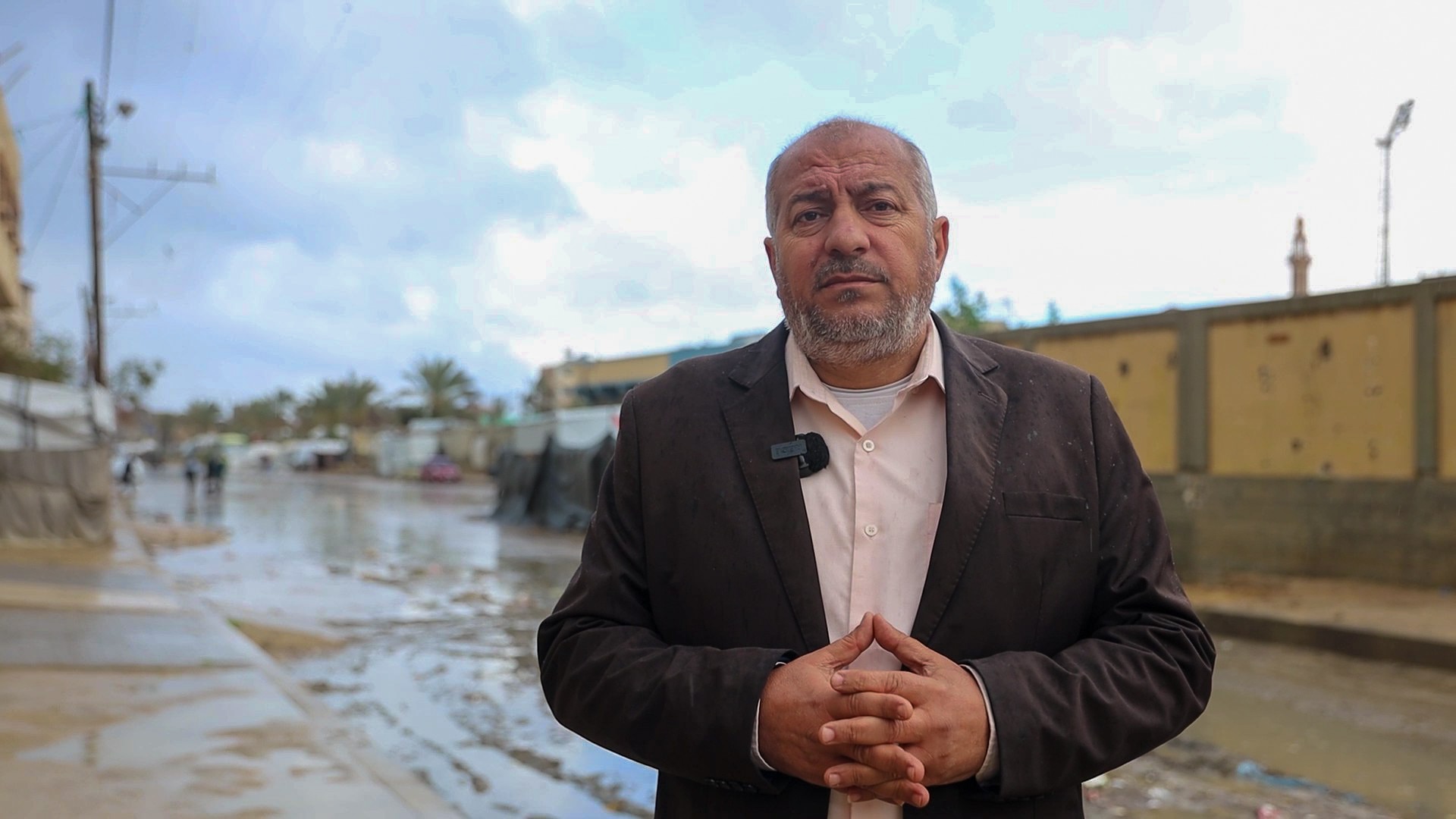 a man wearing a suit jacket stands outside in the rain in front of a muddy and watery road.