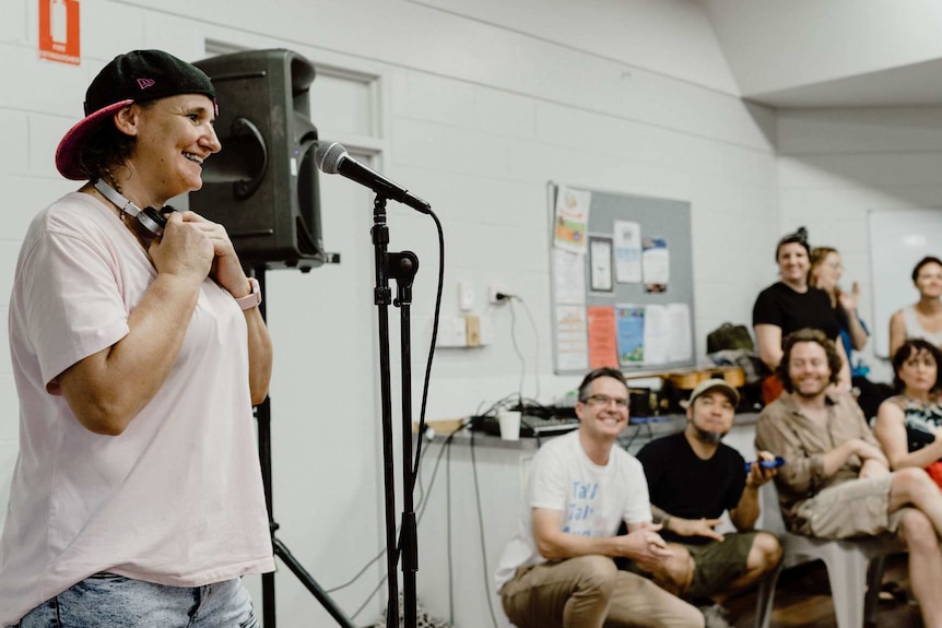 A woman smiles in front of a microphone on a stand, while a crowd looks on.