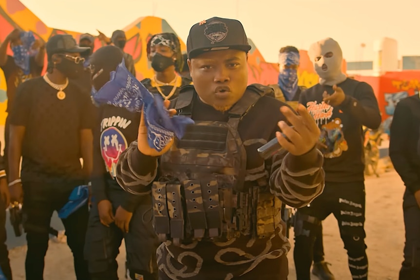 Johnson André appears in a rap music video wearing a bullet proof vest and surrounded by men in masks and jewellery