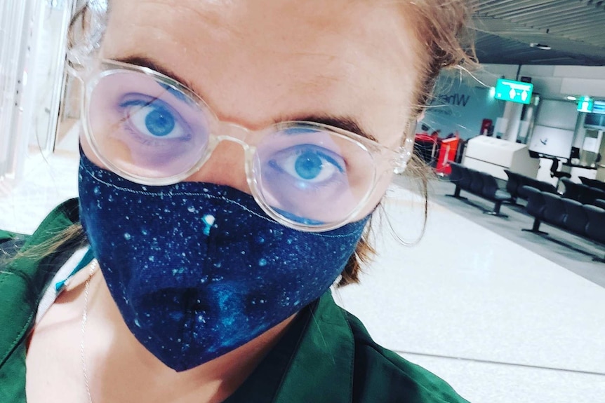 A selfie shot of a woman with spectacles and a blue facemask on in an airport terminal.