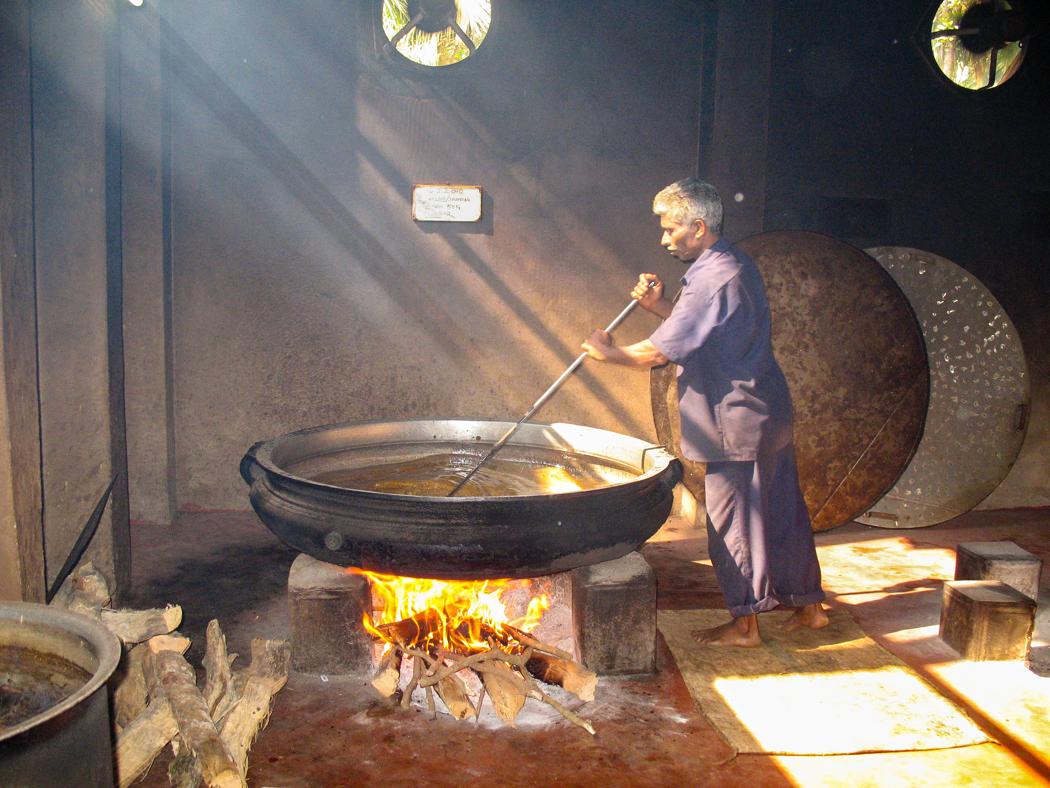 Un hombre indio con cabello blanco y ralo sostiene una gran paleta de acero y se encuentra junto a una tina grande y poco profunda de aceite sobre una llama abierta.