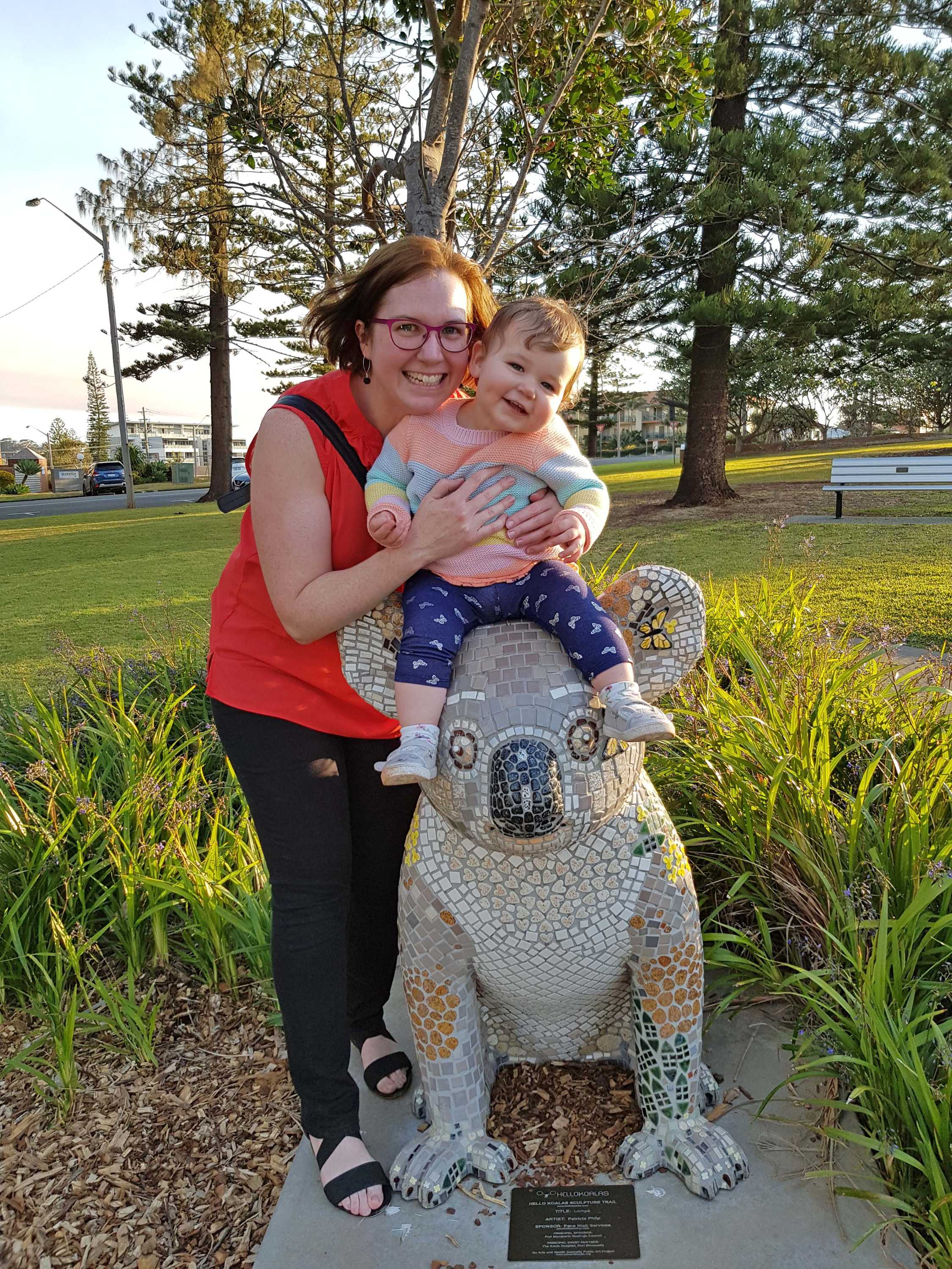 A woman wearing a red singlet holds her baby daughter on top of a koala statue at a park. Both her and baby are smiling broadly.