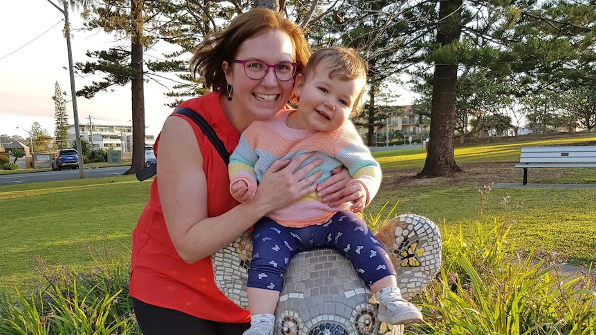 A woman wearing a red singlet holds her baby daughter on top of a koala statue at a park. Both her and baby are smiling broadly.