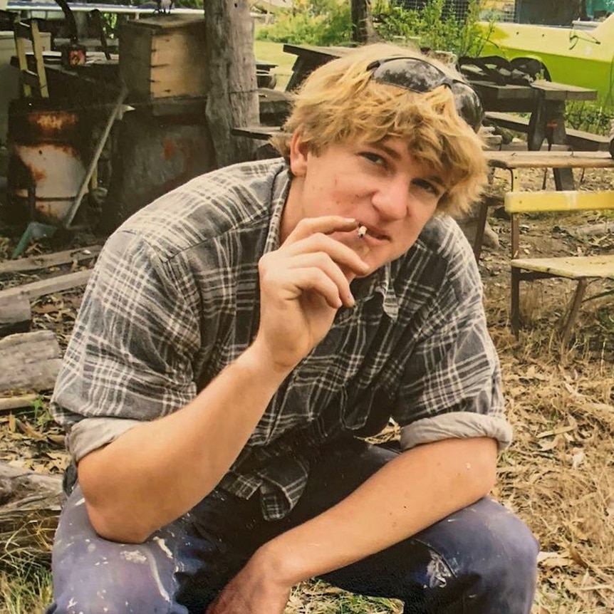 Jack Gerdes sitting on a tree stump is smiling and smoking a cigarette with sunglasses propped up on his forehead.