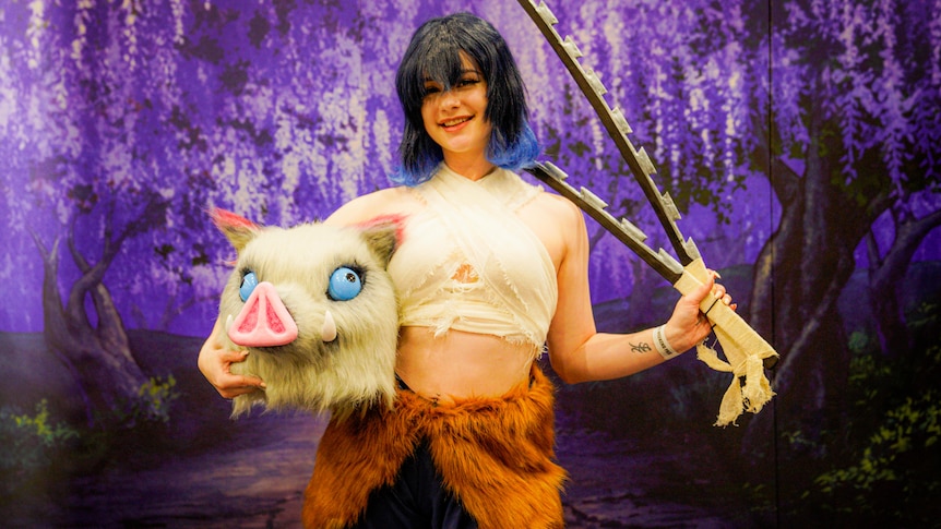 A woman with black and blue hair holding a couple of swords and carrying a cosplay mask