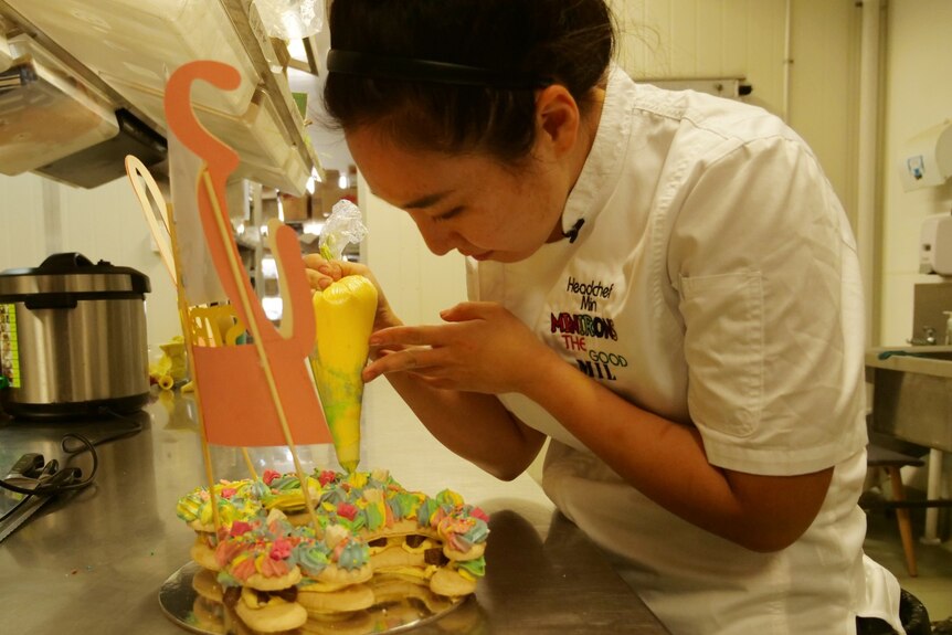 A woman in a commercial kitchen in white chef's uniform piping icing onto a cake