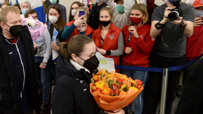 A Russian figure skater walks through an airport wearing a mask, while fans applaud and reporters ask questions as she passes.