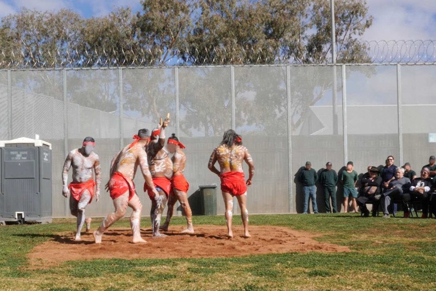 Five men, painted with white markings, in red loincloth and bandannas, dance before a small crowd in a prison yard.