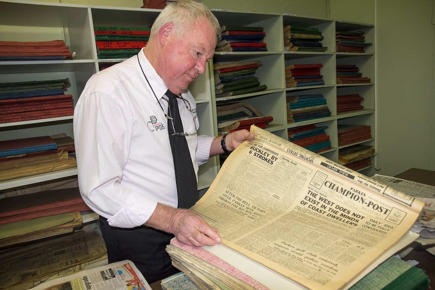 A man looking down at an old newspaper with cupboards full of old editions behind him