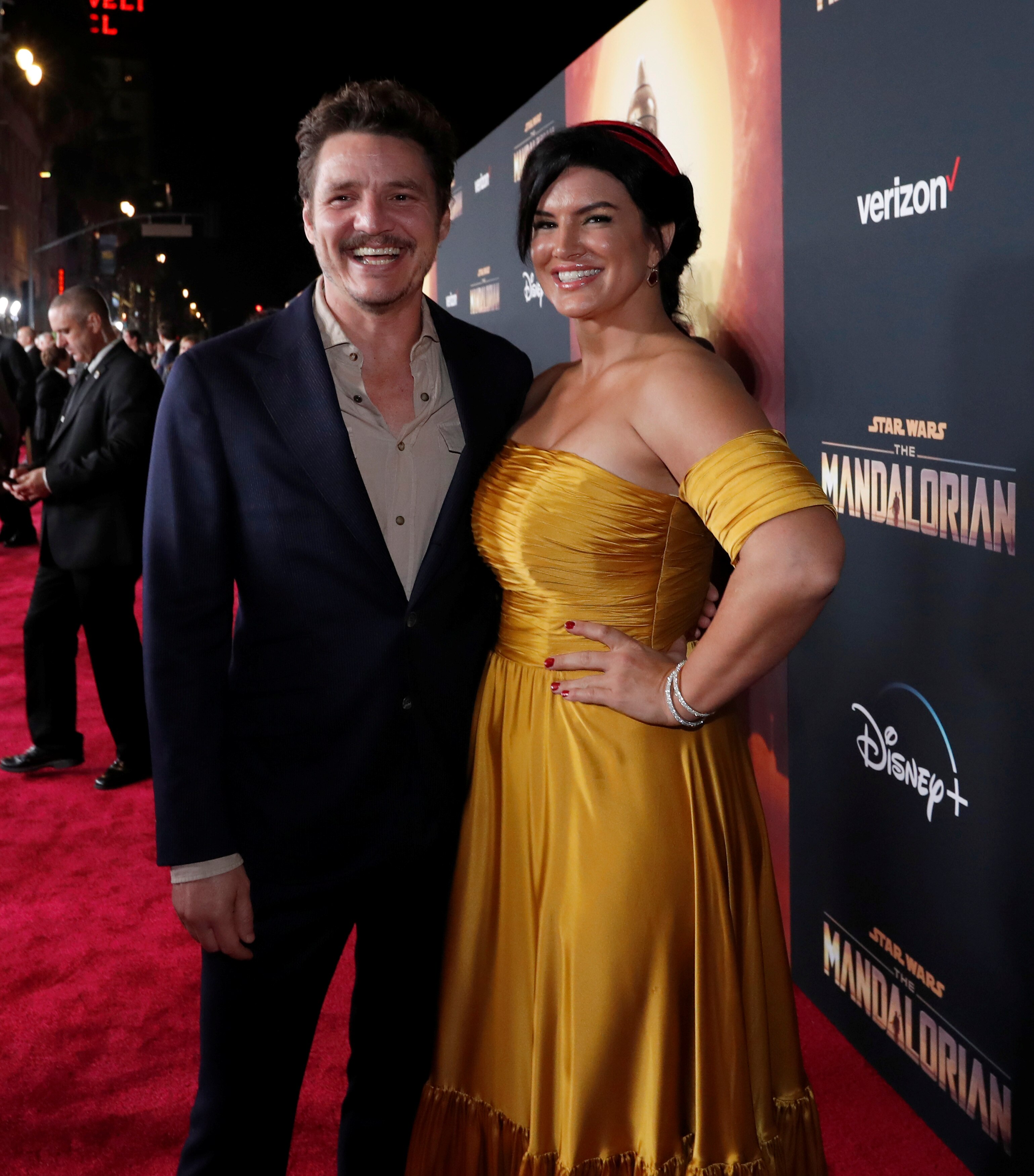 A man in a black suit stands next to a woman in a gold dress on a red carpet