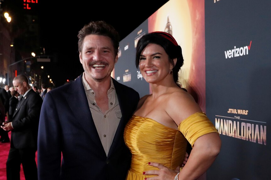 A man in a black suit stands next to a woman in a gold dress on a red carpet