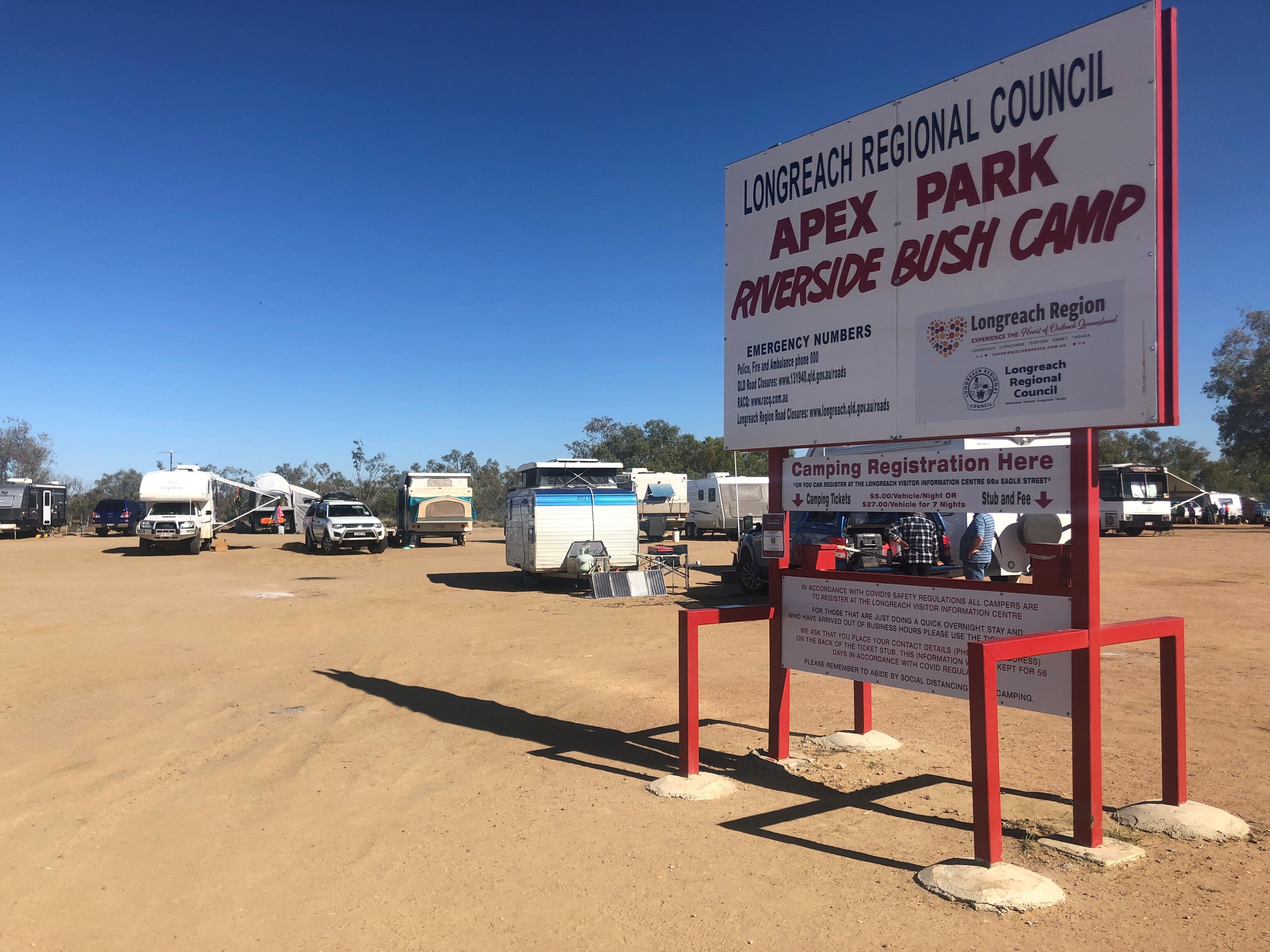 A sign saying apex riverside bush camp Longreach with caravans behind it.