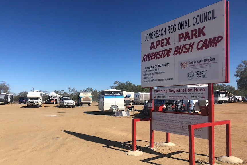 A sign saying apex riverside bush camp Longreach with caravans behind it.