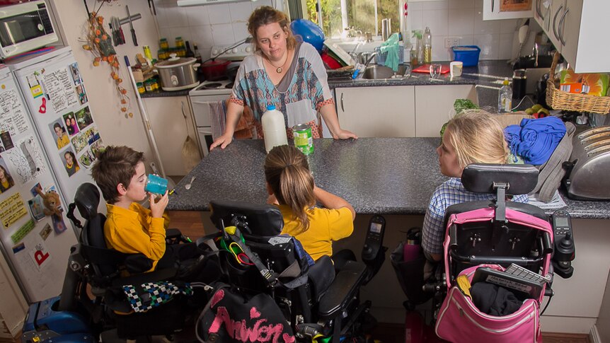 A boy and two girls in power wheelchairs sit at the bench in a kitchen drinking chocolate milk while a woman looks on