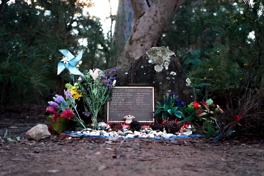 Flowers and gifts laid in front of a plaque on a memorial in a forest.