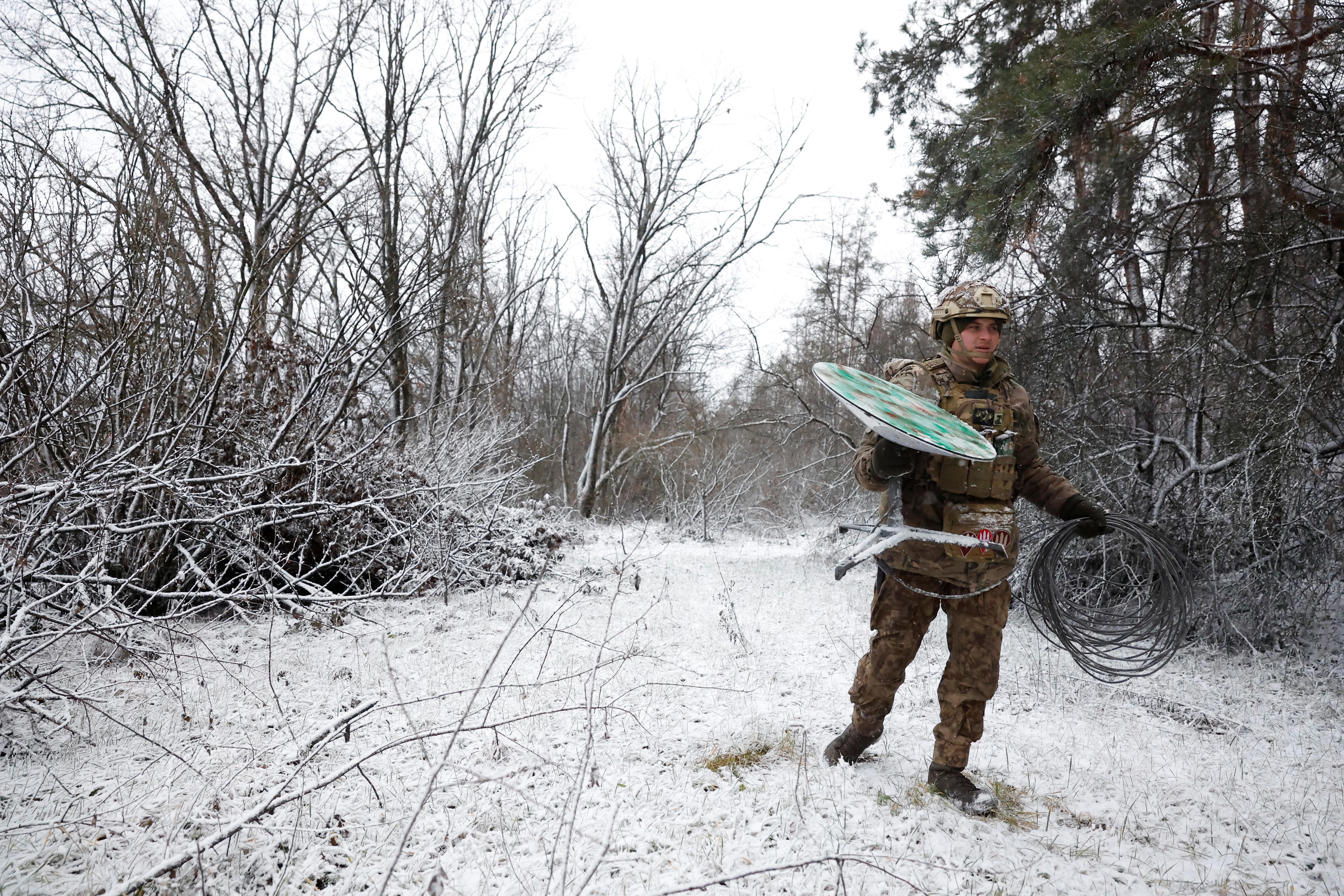 Man in military uniform carries satellite through snow.